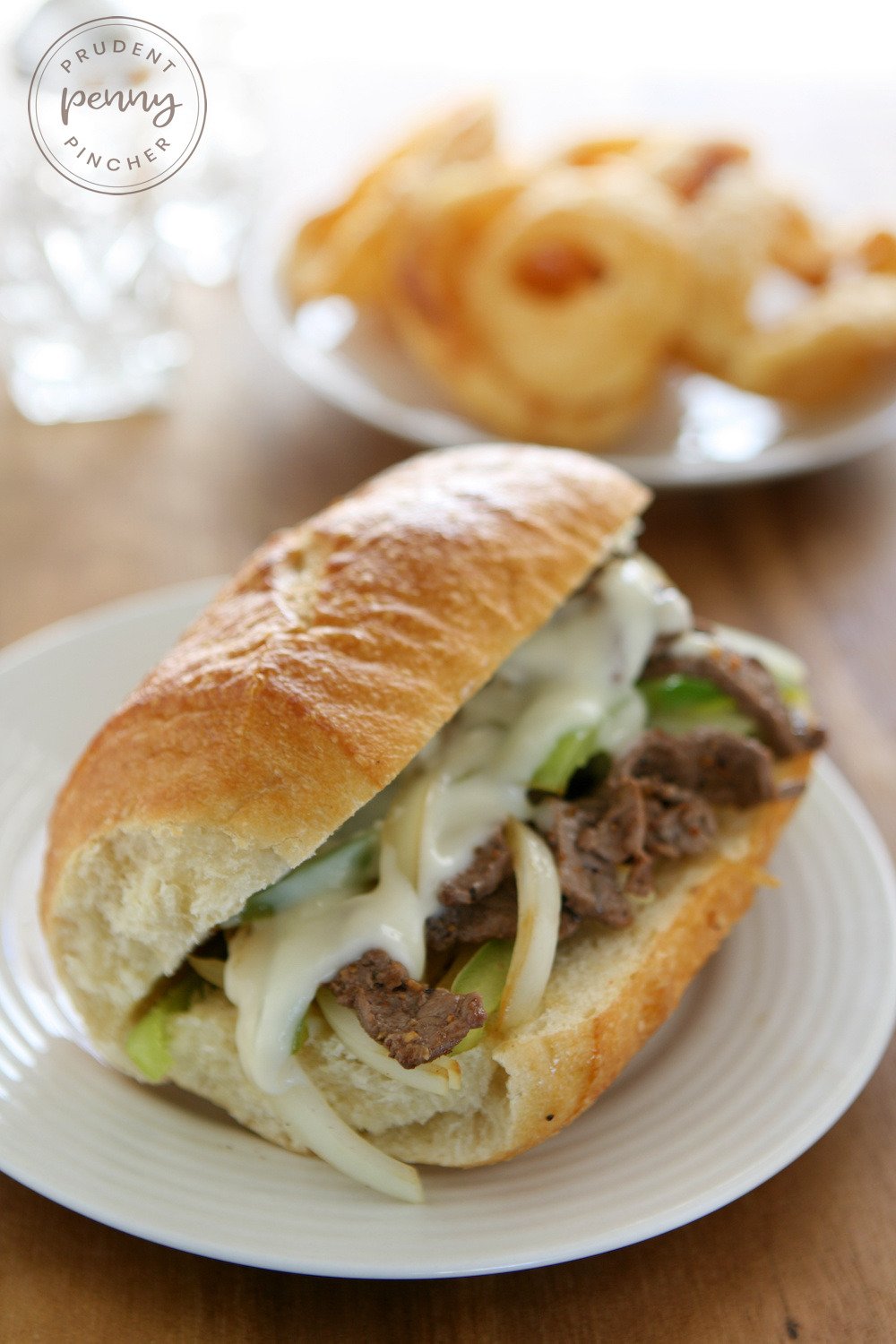 vertical image of Philly cheesesteaks on a plate with a side of onion rings in the background