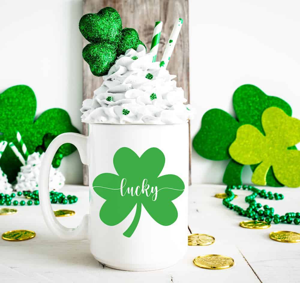 White mug with green shamrock surrounded by glitter shamrocks, gold coins, and whipped cream topping