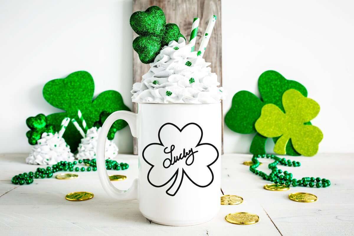 White mug with black outline shamrock surrounded by green glitter shamrocks and gold coins