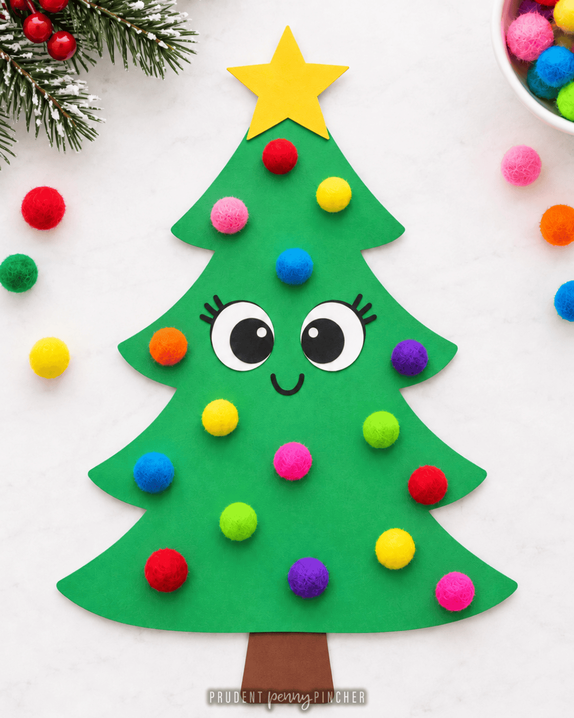 Green paper Christmas tree craft decorated with colorful pom-pom ornaments, a yellow star, and a cute smiling face.