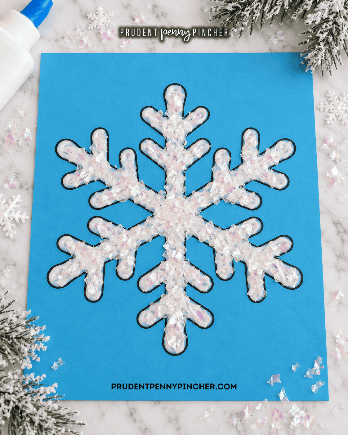 Six-pointed snowflake craft made with white glue and fake snow on blue cardstock, surrounded by winter decorations.